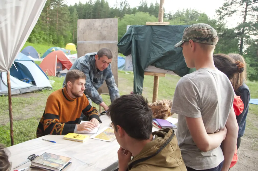 Группа школьников с преподавателем у стола на геологическом слёте, на заднем плане палаточный лагерь