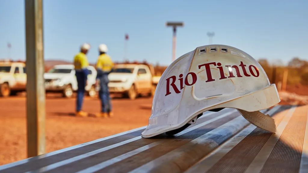 Белая защитная каска с логотипом Rio Tinto лежит на металлической поверхности на промышленной площадке, на заднем плане — двое работников в касках и припаркованные автомобили